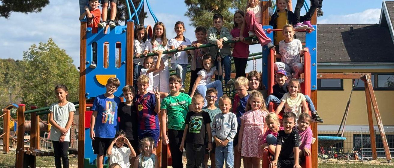 Eine Gruppe von Kindern posiert für ein Foto auf einem Spielplatz, einige klettern auf die Spielgeräte. Der Spielplatz ist von Bäumen und Gras umgeben.