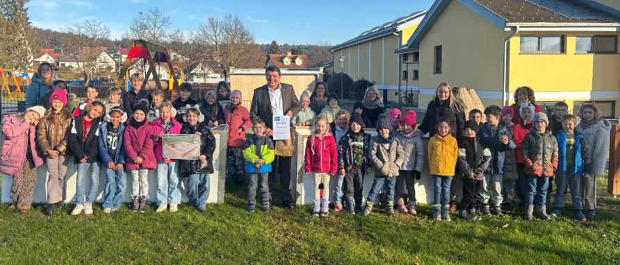 Eine Gruppe von Kindern und Erwachsenen posiert für ein Foto in einem Grasbereich vor einem gelben Gebäude, wobei ein erwachsener Mann ein Zertifikat hält.