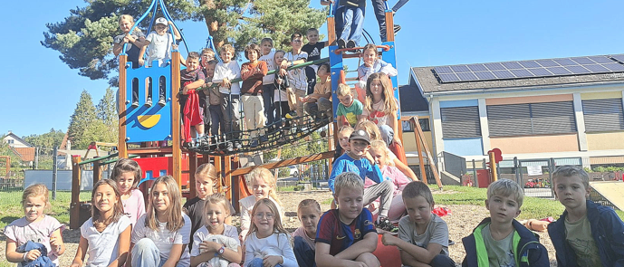Eine Gruppe von Kindern und Jugendlichen versammelt sich um einen Spielplatz mit einem Baum und Solarpanelen im Hintergrund. Einige sitzen auf dem Boden, während andere auf dem Spielgerät stehen.