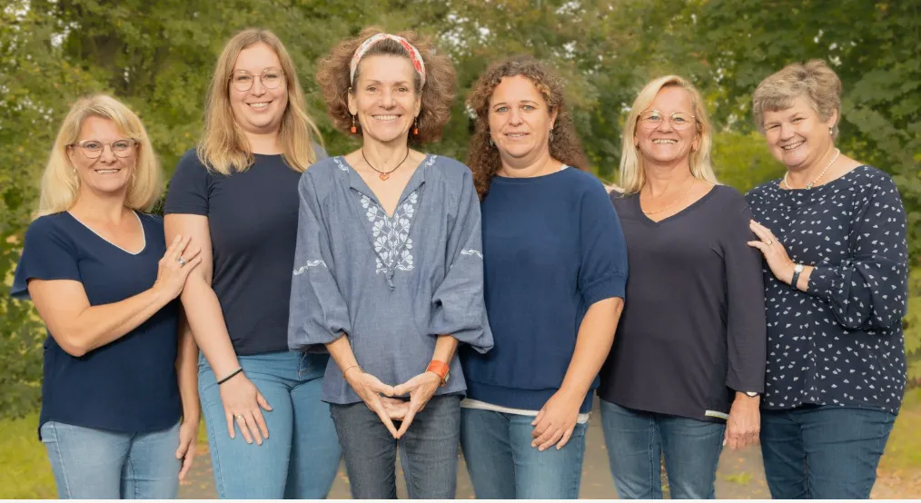 Sechs Frauen in dunkelfarbenen Oberteilen und Jeans lächeln und posieren für ein Gruppenfoto in einem grünen, belaubten Park.