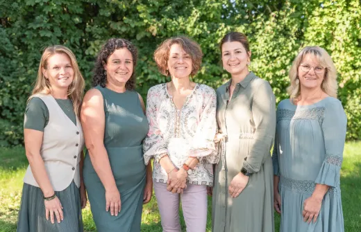 Fünf Frauen stehen in einer Reihe in einem Garten, lächeln und posieren für ein Foto.