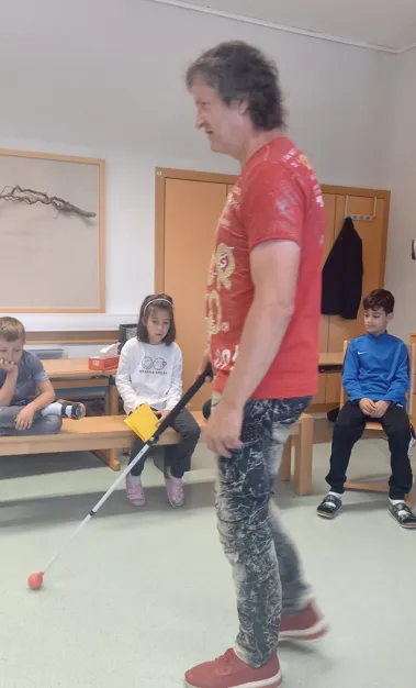 Ein Mann in einem roten T-Shirt benutzt einen Stock, um einen Raum mit Kindern zu navigieren, die auf Bänken sitzen.