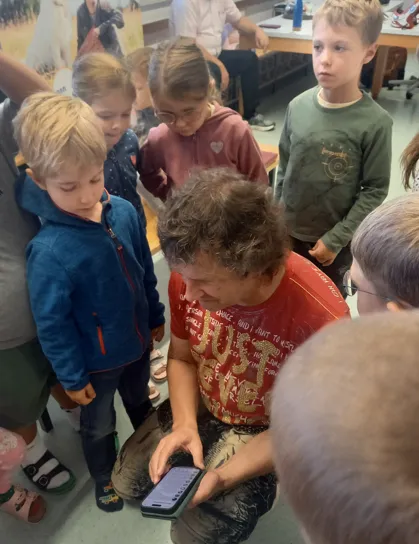 Eine Gruppe von Kindern und einem Erwachsenen versammelt sich um einen Mann, der auf sein Handy schaut. Die Kinder stehen und beobachten ihn.
