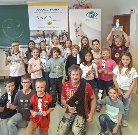 Eine Gruppe von Kindern und einer erwachsenen Frau posiert für ein Foto in einem Klassenzimmer. Sie halten Karten hoch und lächeln. Hinter ihnen ist ein Banner mit dem Text 'Augen auf Pfoten' angebracht.