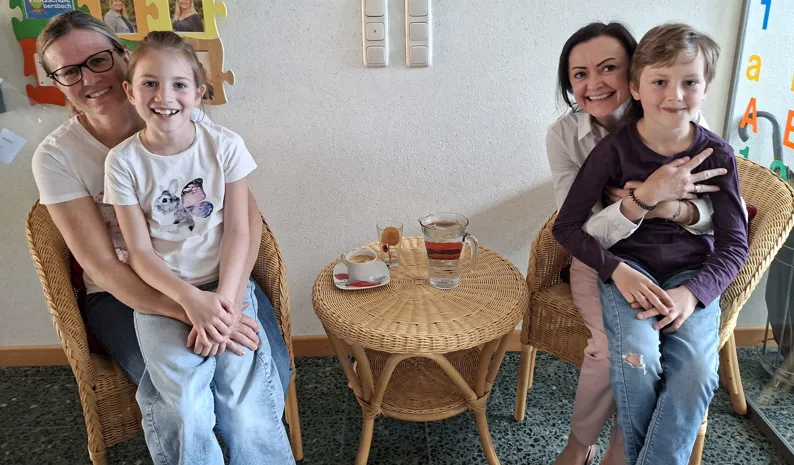 Drei Frauen und zwei Kinder sitzen auf Stühlen und lächeln für ein Foto. Ein Tisch mit einer Tasse und einer Karaffe in der Mitte. Eine Wand mit Buchstaben und Bildern hinter ihnen.