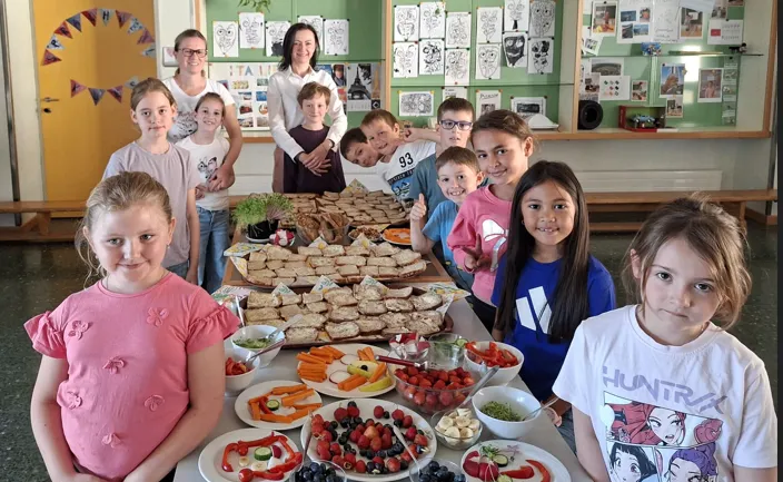 Eine Gruppe von Kindern und Erwachsenen posiert für ein Foto in einem Klassenzimmer mit einem Tisch voller Sandwiches, Obst und Gemüse.
