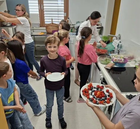 Eine Gruppe von Kindern befindet sich in der Küche. Ein Junge hält einen Teller mit Obst. Einige Kinder betrachten das Essen, während andere herumstehen.