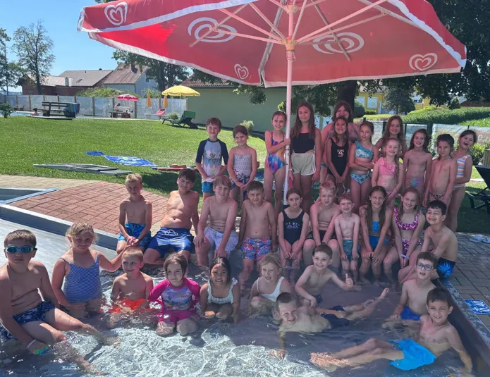 Eine Gruppe von Kindern in Badeanzügen im Pool, mit einem Schirm und Erwachsenen um sie herum, an einem sonnigen Tag.