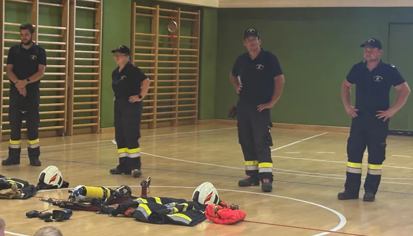 Vier Feuerwehrleute in Uniform stehen auf einem Holzboden mit Feuerwehrausrüstung, Helmen und einem Feuerlöscher. Eine Frau schaut in die Kamera. Hinter ihnen sind Holzkletterstangen an der grünen Wand befestigt.