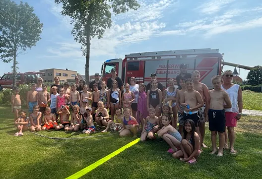 Eine Gruppe von Kindern und Erwachsenen posiert an einem sonnigen Tag für ein Foto. Ein Feuerwehrauto ist im Hintergrund.
