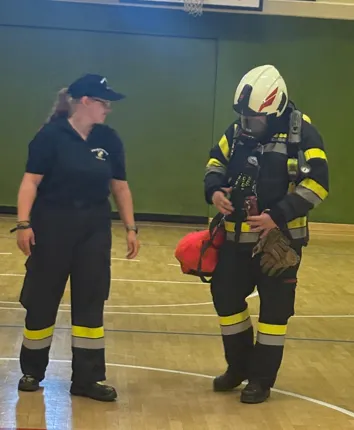 Eine Frau und ein Feuerwehrmann in voller Ausrüstung stehen auf einem Holzboden in einer Halle. Der Feuerwehrmann hält eine rote Tasche und ein Werkzeug.