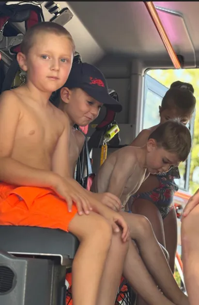 Vier Jungen sitzen in einem Fahrzeug, möglicherweise einem Feuerwehrauto. Einer trägt ein orangefarbenes Shirt, ein anderer eine Mütze, und zwei sind unbekleidet. Eines Mädchen in einem Blumenbadeanzug steht hinter ihnen.