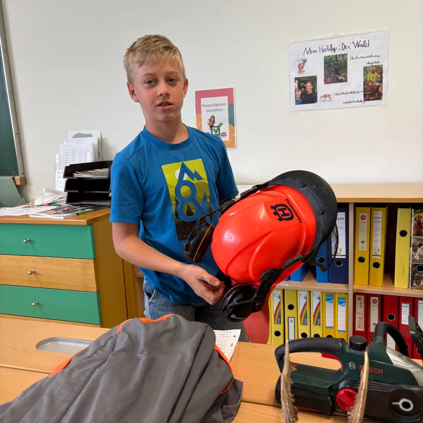 Ein Junge hält einen roten Helm und Schutzbrille in einem Klassenzimmer. Verschiedene Werkzeuge und Ausrüstung sind auf dem Schreibtisch.