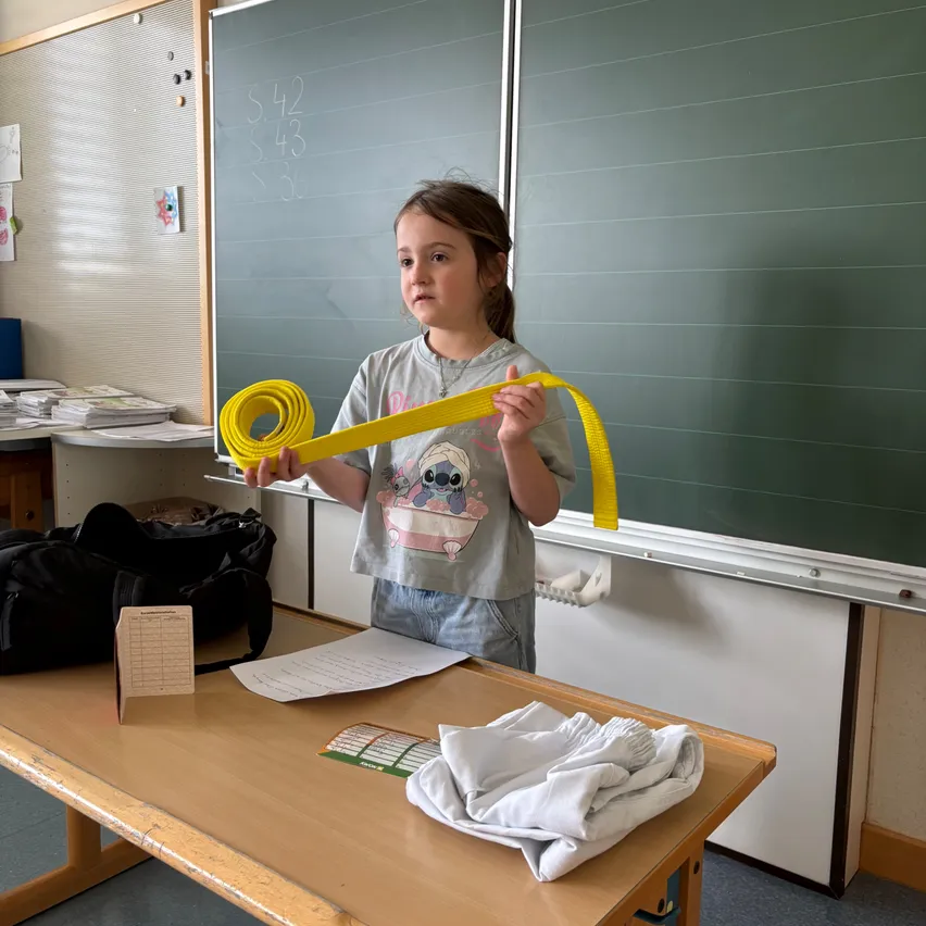 Ein junges Mädchen hält ein gelbes Band vor einem Klassenzimmer. Sie steht an einem Schreibtisch mit einem Blatt Papier und einigen Kleidungsstücken darauf.