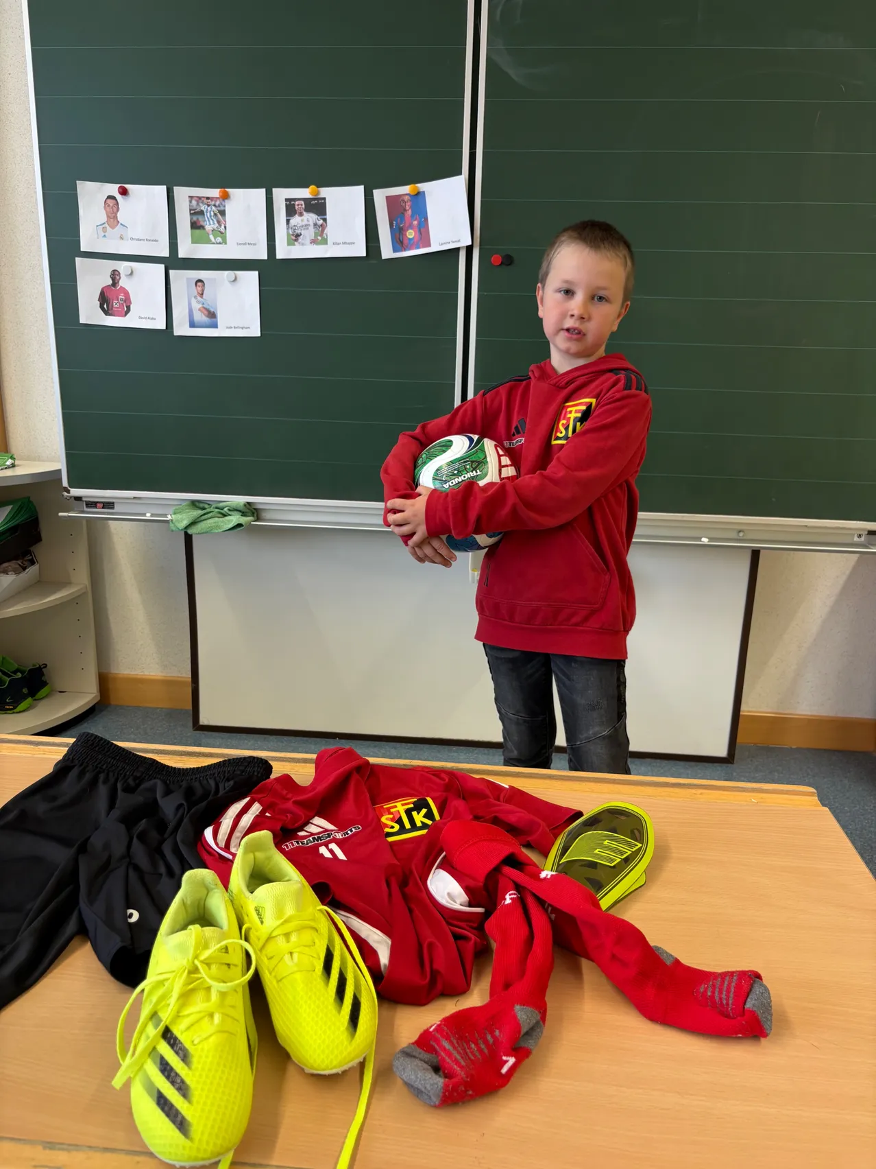 Ein junger Junge in einem roten Pullover steht vor einem Klassenzimmer und hält einen Fußball. Dahinter hängt eine Tafel mit angehefteten Fotos. Auf dem Tisch davor befinden sich Sportkleidung und Schuhe.