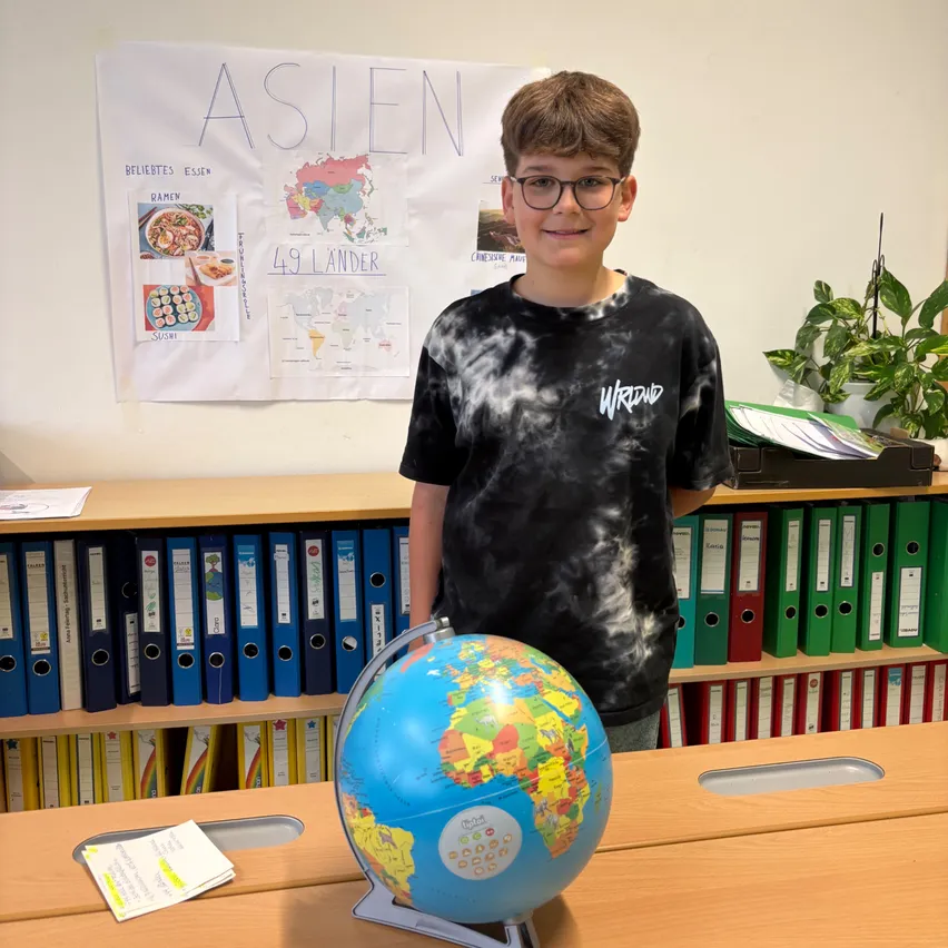 Ein junger Junge steht in einem Klassenzimmer neben einem Globus, trägt eine Brille und ein schwarzes T-Shirt. Hinter ihm zeigt eine weiße Tafel eine Karte und eine Liste von Ländern.