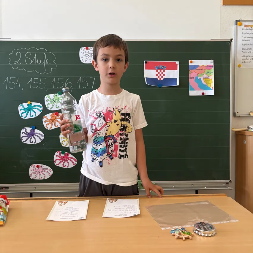Ein junger Junge steht vor einem Klassenzimmer-Schwarzbrett, hält eine Flasche und lächelt, mit verschiedenen Zeichnungen und einer Flagge hinter ihm.