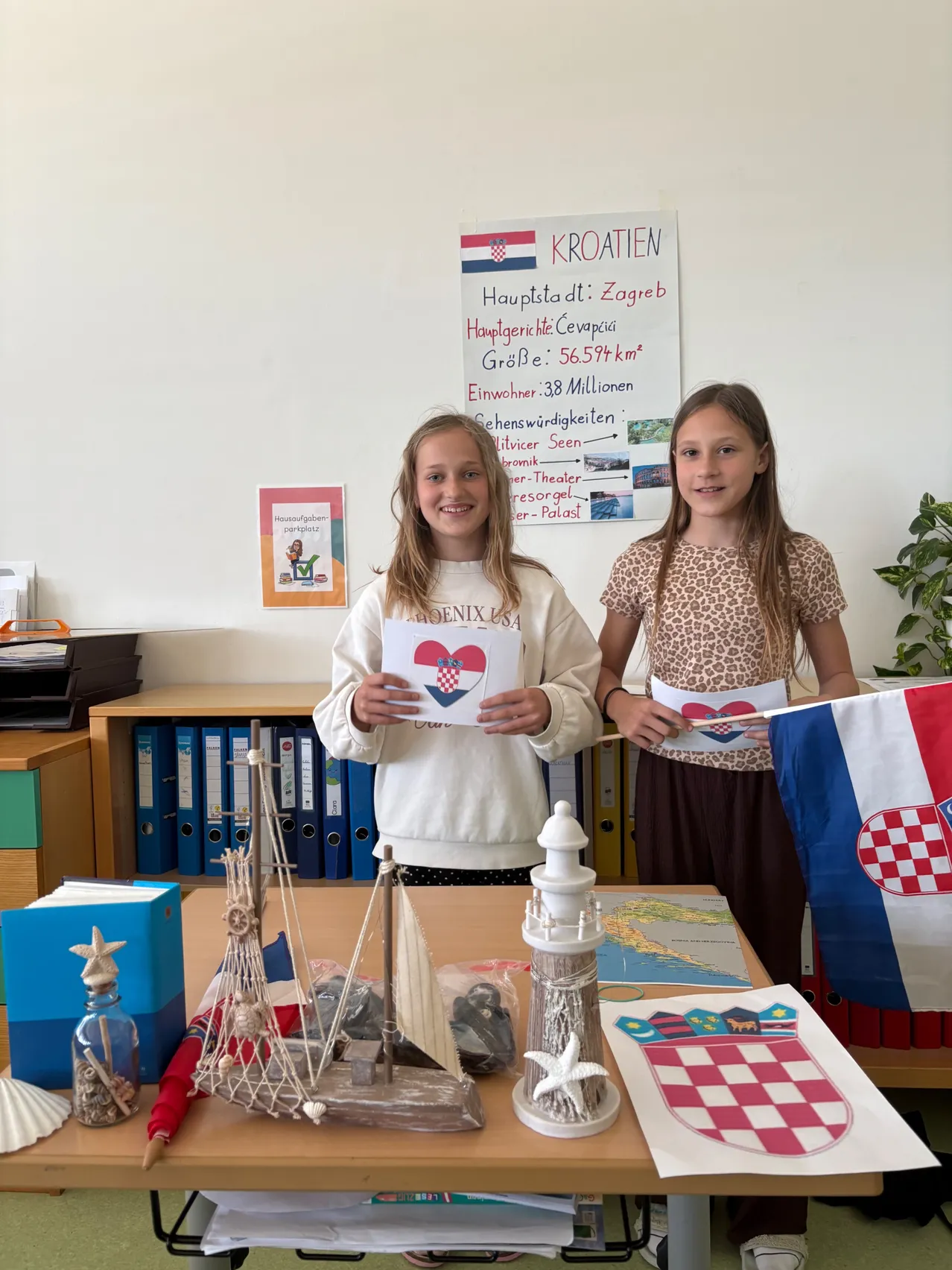 Zwei Mädchen stehen in einem Klassenzimmer, eines hält eine Karte mit einer kroatischen Flagge und das andere eine Flagge. Hinter ihnen steht ein Schreibtisch mit verschiedenen Gegenständen, einem Poster mit Informationen über Kroatien und einem Topf.