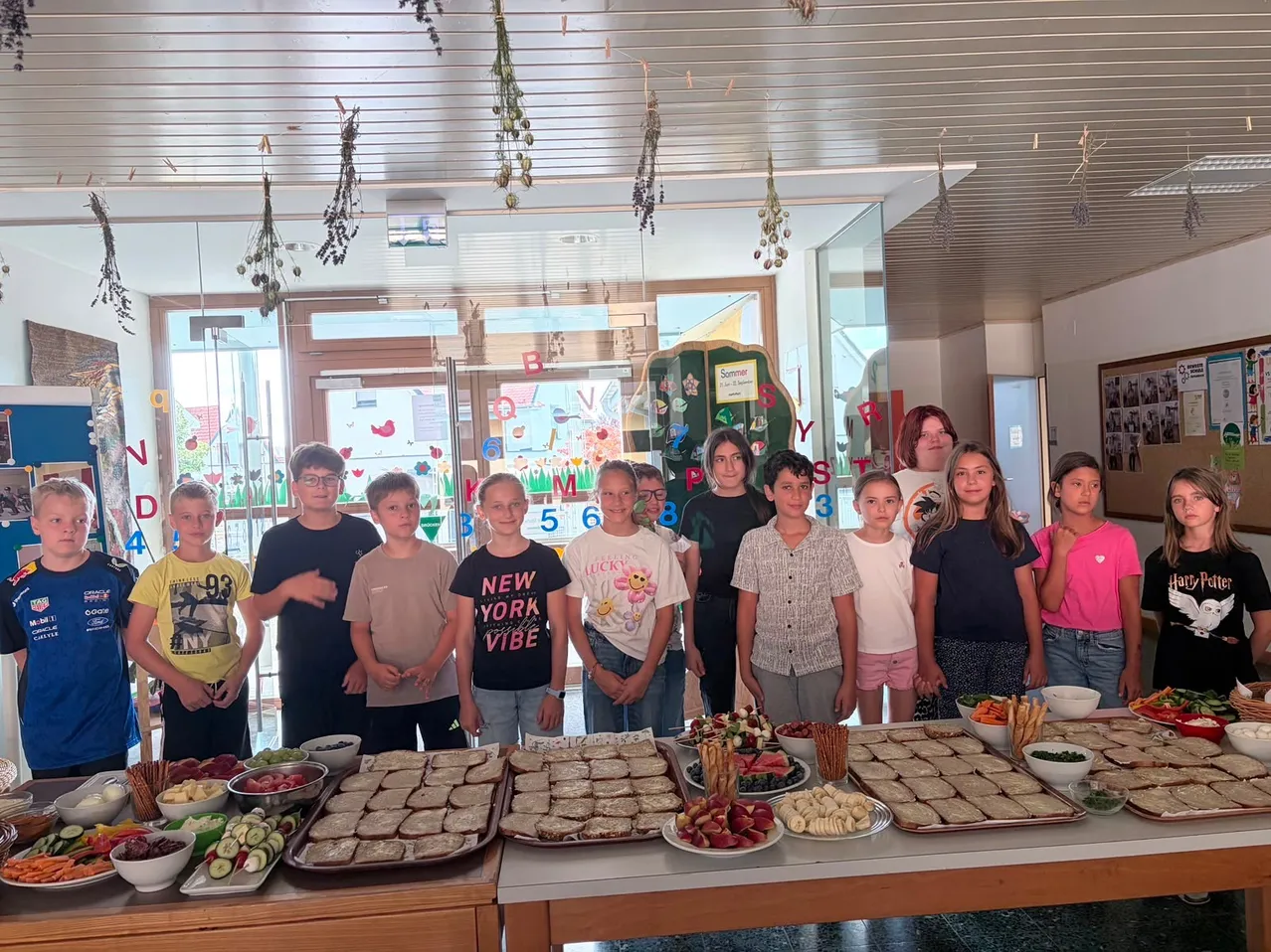 Eine Gruppe von Kindern und Erwachsenen steht lächelnd vor einem Buffettisch mit Sandwiches, Obst und Gemüse. Der Raum hat eine Glastür, Pflanzen und eine bunte Wand.