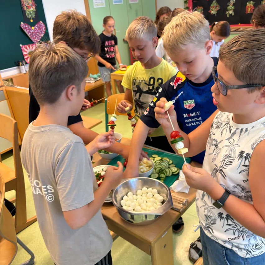 Mehrere Jungen in einem Klassenzimmer bereiten Essen auf Stöcken vor. Ein Junge hat eine Tomate am Stiel im Mund.