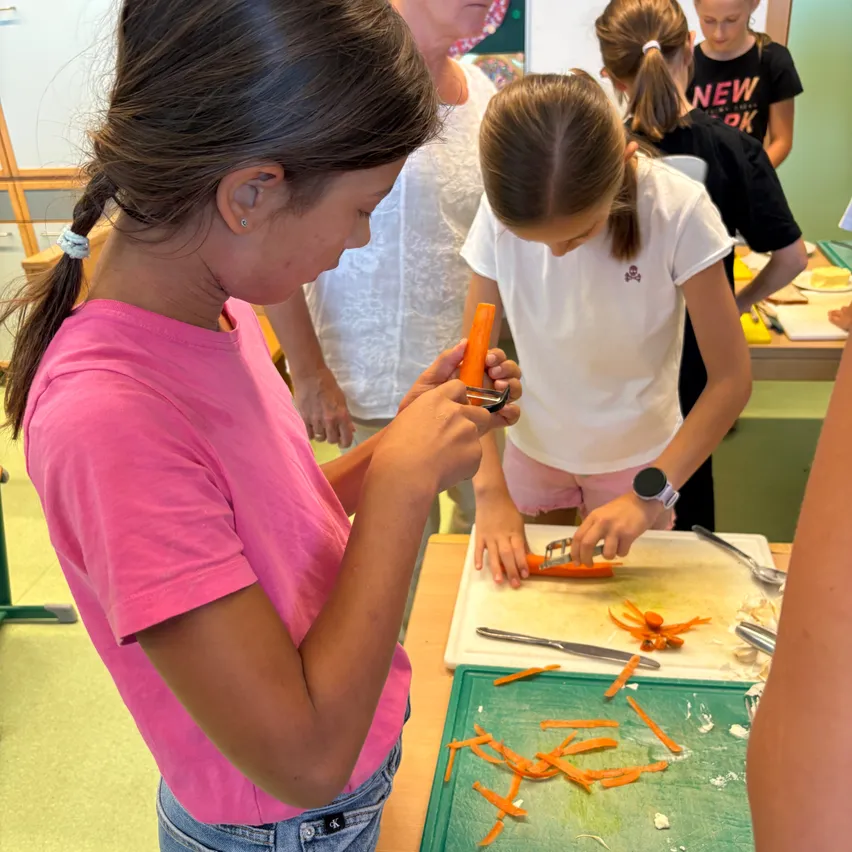 Zwei Mädchen in einer Küche, eine in einem rosa T-Shirt schält eine Karotte, während eine andere in einem weißen T-Shirt Karotten auf einem grünen Schneidebrett schneidet. Andere Kinder und Erwachsene sind im Hintergrund.