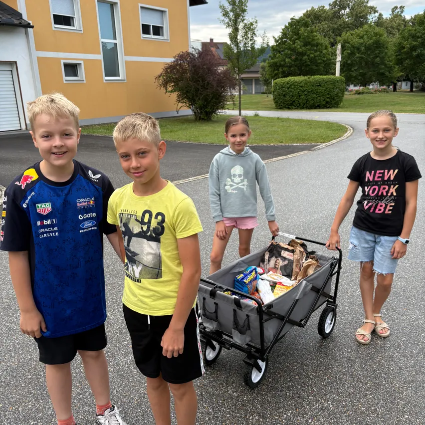 Vier Kinder stehen auf einer Einfahrt. Ein Kind hält einen Wagen voller Lebensmittel. Sie alle lächeln. Hinter ihnen steht ein Haus und einige Bäume.