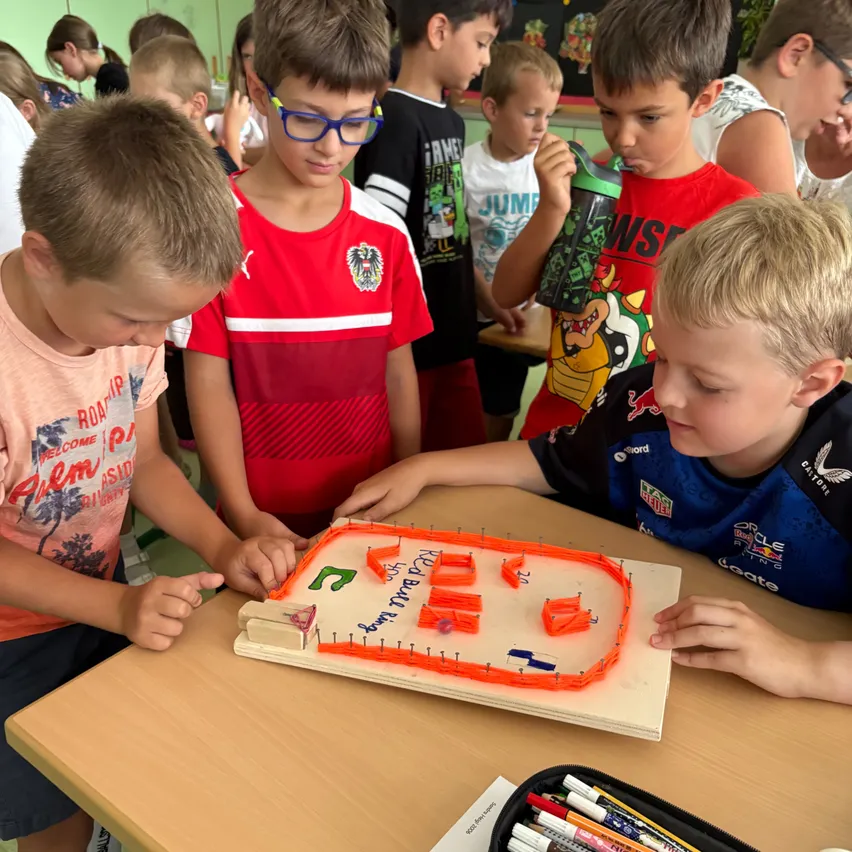 Mehrere Jungen sind um einen Tisch versammelt und spielen ein Spiel mit einer orangefarbenen Strecke. Ein Junge zeigt und ein anderer hält einen Stift.
