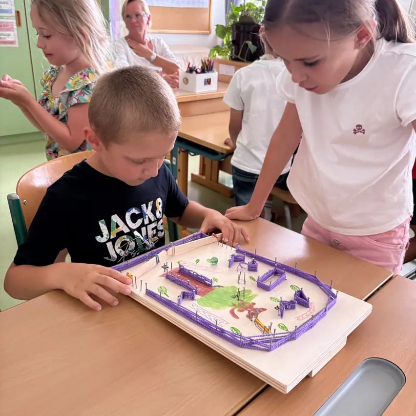Ein Junge und ein Mädchen konzentrieren sich auf ein Brettspiel in einem Klassenzimmer, während andere Kinder und eine Lehrerin in der Nähe sind.