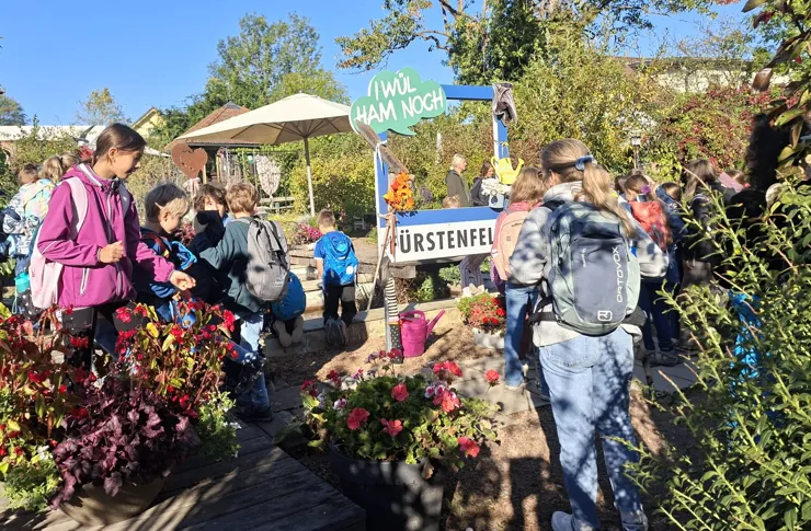 Eine Gruppe von Kindern und Erwachsenen versammelt sich um einen blau-weißen Stand mit einem Schild, das 'Würstenfel' besagt. Sie befinden sich in einem Garten mit verschiedenen Pflanzen und Blumen.