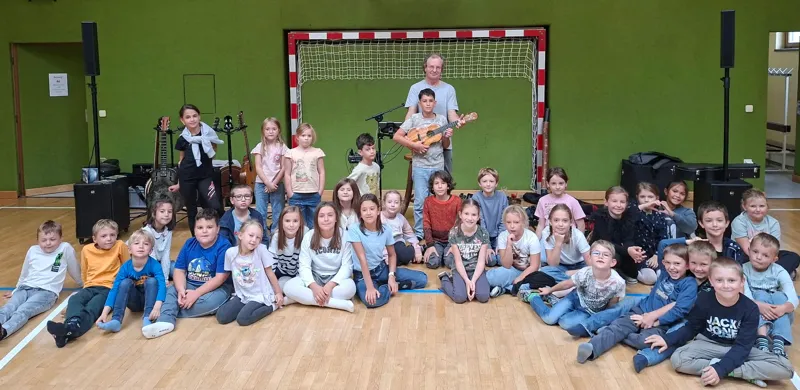 Eine Gruppe von Kindern und zwei Erwachsenen versammelt sich in einem Raum. Die Kinder sitzen auf dem Boden und die Erwachsenen stehen hinter ihnen. Einer der Erwachsenen hält eine Gitarre und scheint den Kindern etwas beizubringen.