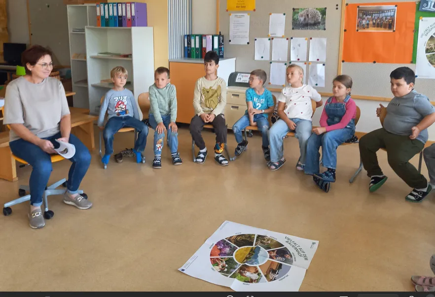 Eine Gruppe von Kindern und einer erwachsenen Frau sitzen im Kreis auf Stühlen in einem Klassenzimmer.