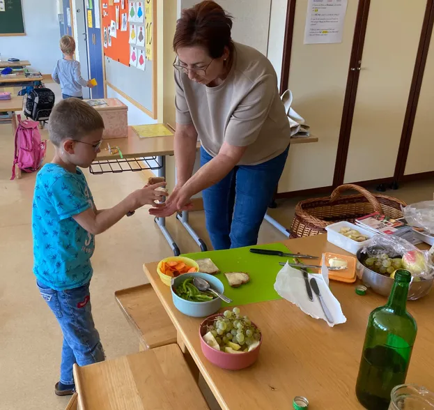 Eine Frau hilft einem kleinen Jungen, ein Sandwich mit Gemüse auf einem Tisch in einem Klassenzimmer zuzubereiten.