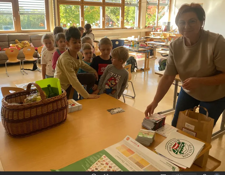 Eine Gruppe von Kindern und eine erwachsene Frau stehen um einen Tisch in einem Klassenzimmer. Der Tisch hat einen Korb, Bücher und eine Tasche darauf. Die Frau lächelt und schaut die Kinder an.