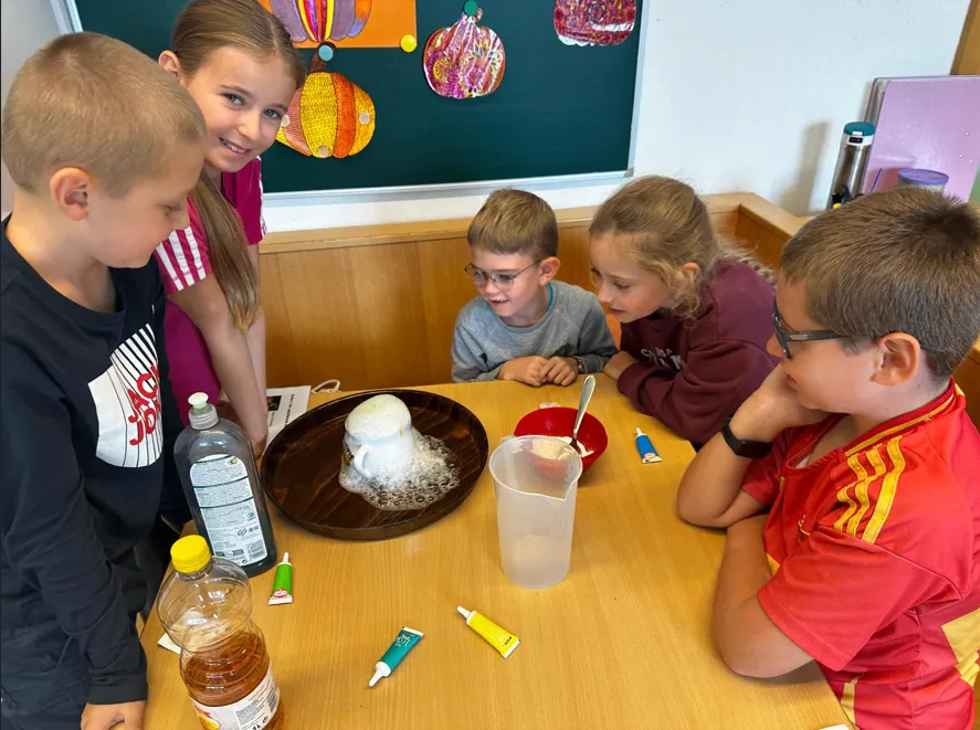 Eine Gruppe von Kindern ist um einen Tisch versammelt und beobachtet ein wissenschaftliches Experiment. Ein Vulkanmodell steht in der Mitte, mit einer Tasse und einem Teller in der Nähe. Verschiedene Flaschen und Röhren liegen auf dem Tisch, und ein Tafel mit Kürbisdekorationen ist im Hintergrund.