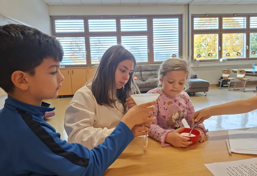 Drei Kinder versammeln sich um einen Tisch in einem Klassenzimmer. Ein Junge hält eine Trichter, ein Mädchen hält eine Schüssel und ein anderes Mädchen schaut sie an.