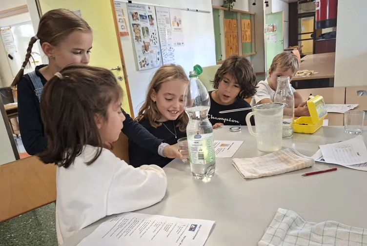 Fünf Kinder sitzen an einem Tisch in einem Klassenzimmer. Sie machen ein wissenschaftliches Experiment mit einer Wasserflasche und einem Messbecher.