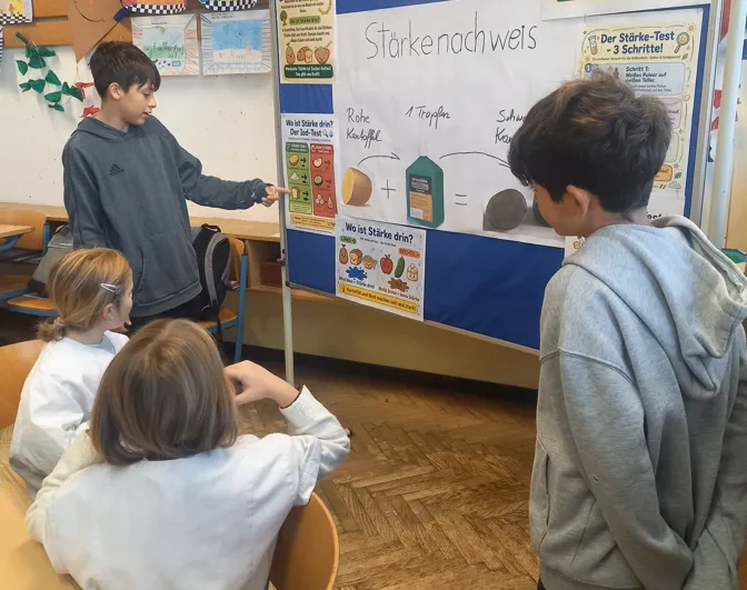 Eine Gruppe von Kindern in einem Klassenzimmer versammelt sich um ein Poster mit einem Diagramm und Text über Stärke.