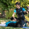 Ein Trainer führt eine Frau in einer Yoga-Pose auf einem Grasfeld. Der Trainer kniet neben der Frau, die auf dem Rücken liegt. Beide tragen Sportbekleidung. Bäume mit grünen Blättern sind im Hintergrund.