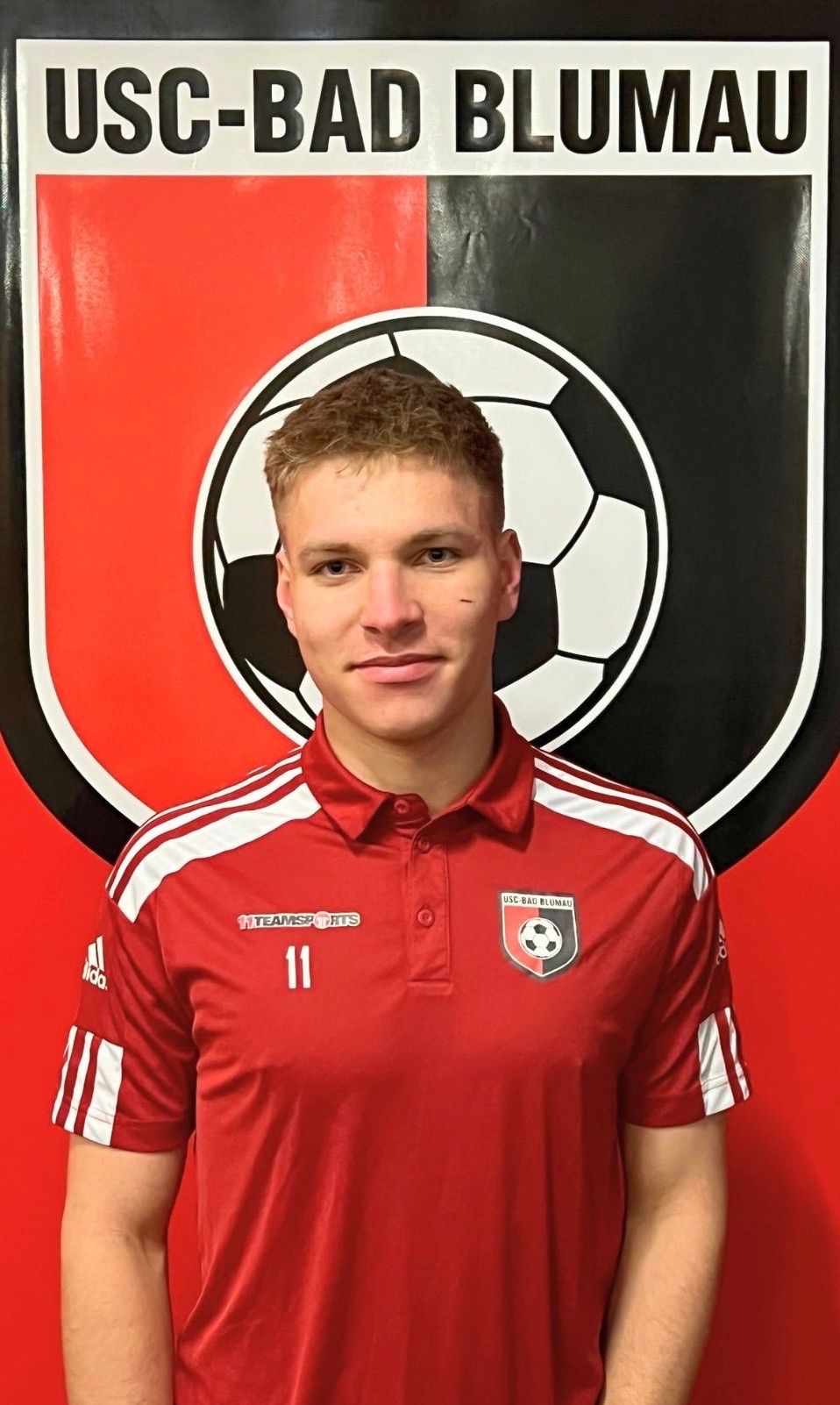 A soccer player in a red jersey with the number 11 poses in front of a red and black backdrop with a soccer ball design.