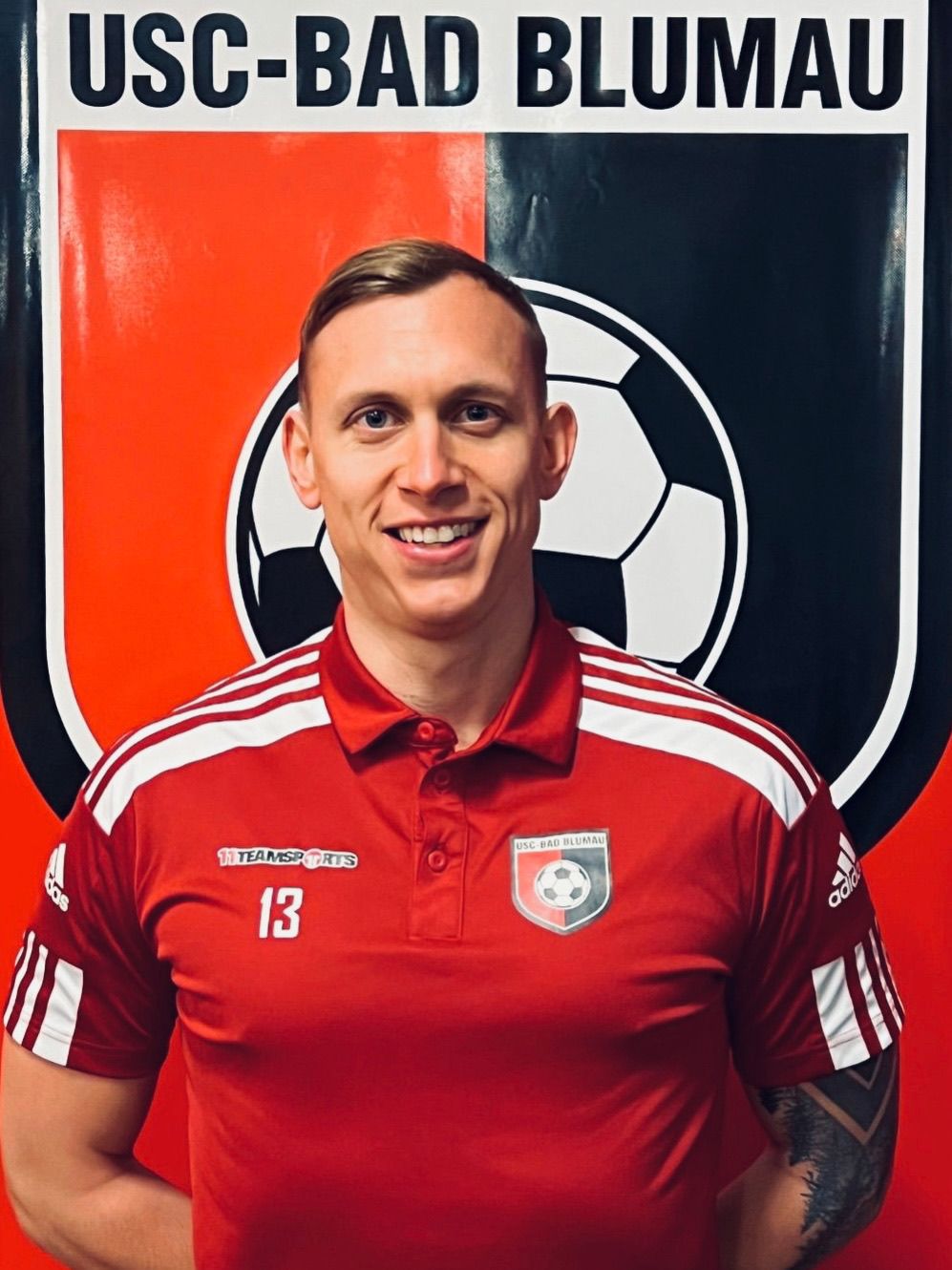 A man wearing a red shirt with the number 13, smiling in front of a red and black backdrop with a soccer ball.