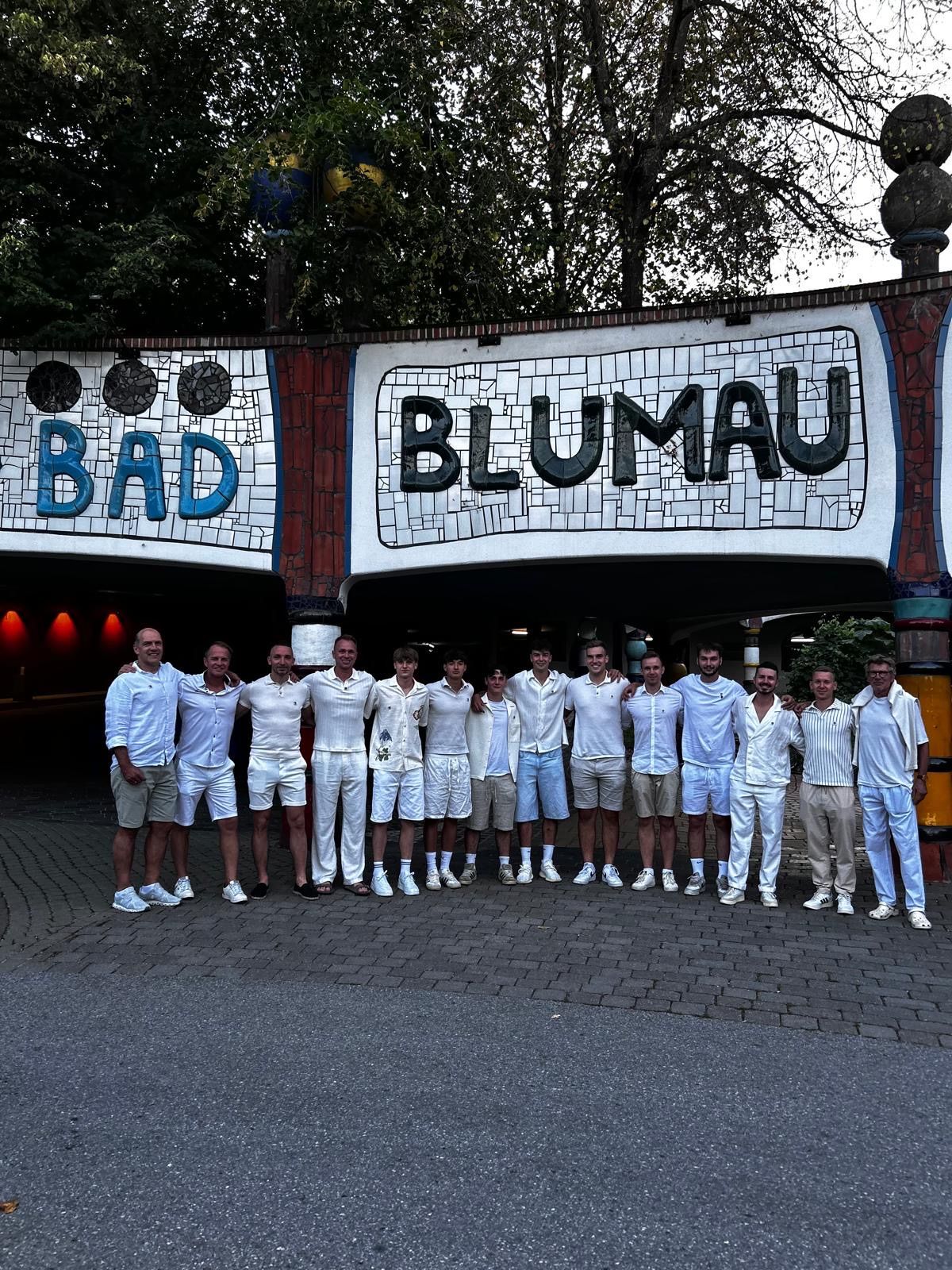 Eine Gruppe von Männern in weißen Outfits posiert für ein Foto vor einem bunten Tor mit den Worten 'BAD' und 'BLUMAU'.
