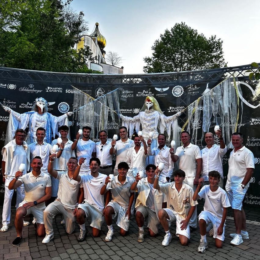 Eine Gruppe von Männern in weißen Outfits posiert für ein Foto vor einem dekorierten Hintergrund mit zwei Personen in weißen Kostümen, die Weingläser halten.