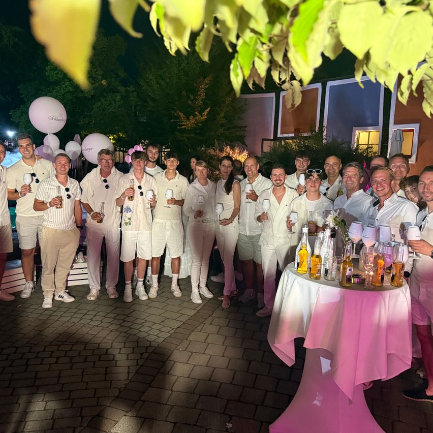 Eine Gruppe von Menschen in weißen Kleidungsstücken posiert nachts für ein Foto, hält Getränke und Ballons. Sie stehen vor einem Gebäude mit farbenfrohen Wänden.