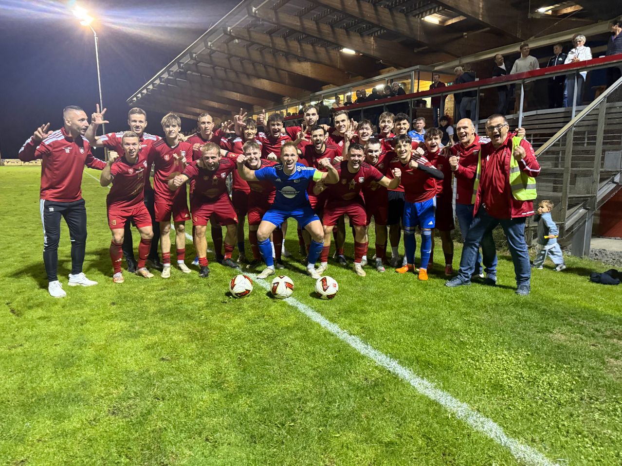 Eine Fußballmannschaft feiert auf dem Feld bei Nacht, die Spieler tragen rote Trikots und ein Trainer eine Warnweste.