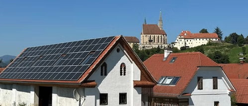 Ein Haus mit Solarpanelen auf dem Dach und einer Kirche im Hintergrund. Das Haus ist weiß mit einem roten Dach.