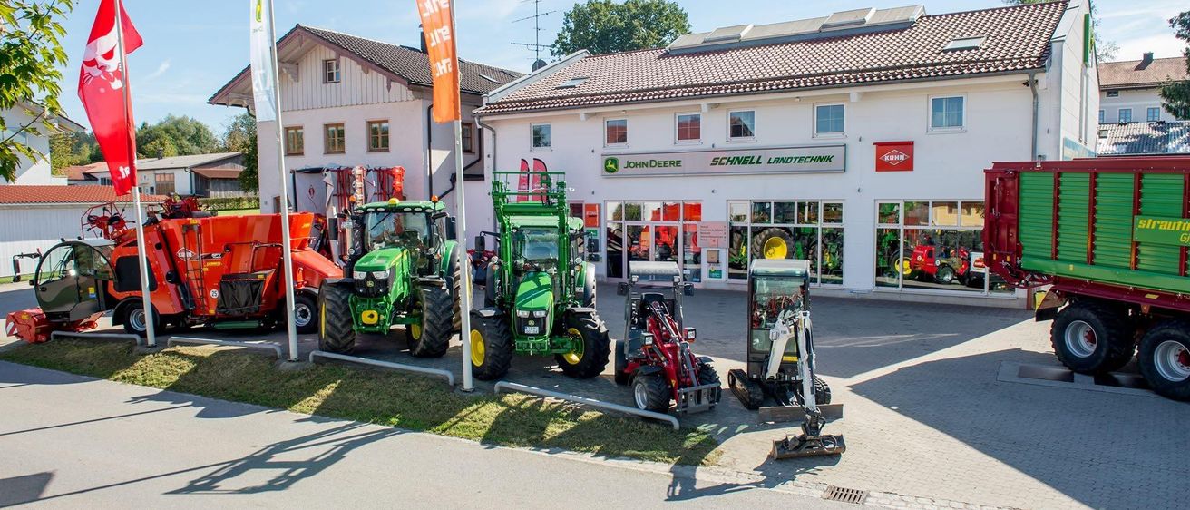 Ein John Deere Händler mit mehreren Traktoren und Maschinen, die an einem sonnigen Tag draußen ausgestellt sind.