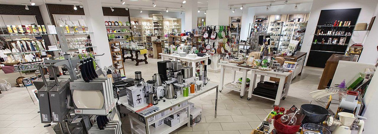 Ein gut beleuchteter Küchenwarenladen mit verschiedenen ausgestellten Artikeln, einschließlich Teller, Töpfe und Kaffeemaschinen. Der Laden hat ein sauberes und organisiertes Layout.