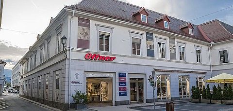 Ein weißes Gebäude mit roten Dächern und einem roten Schild, das 'Offner' steht. Das Gebäude hat mehrere Fenster und eine Glastür. Vorne stehen Topfpflanzen und ein Mülleimer. Rechts steht ein gelber Schirm.