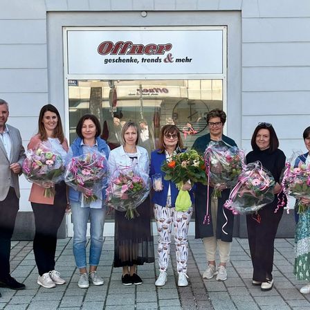Eine Gruppe von Frauen und Männern steht vor einem Geschäft mit einem Schild, das 'Offner Geschenke, Trends & Mehr' besagt. Sie halten Blumensträuße und scheinen für ein Foto zu posieren.