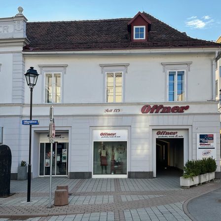 Ein weißes Gebäude mit braunem Dach und rotem Rand hat ein Schild mit der Aufschrift 'Offner'. Das Gebäude hat mehrere Fenster und eine Glastür mit einer Puppe im Inneren. Eine Straßenlampe und ein Schild mit der Aufschrift 'Offnerplatz' befinden sich auf der linken Seite.